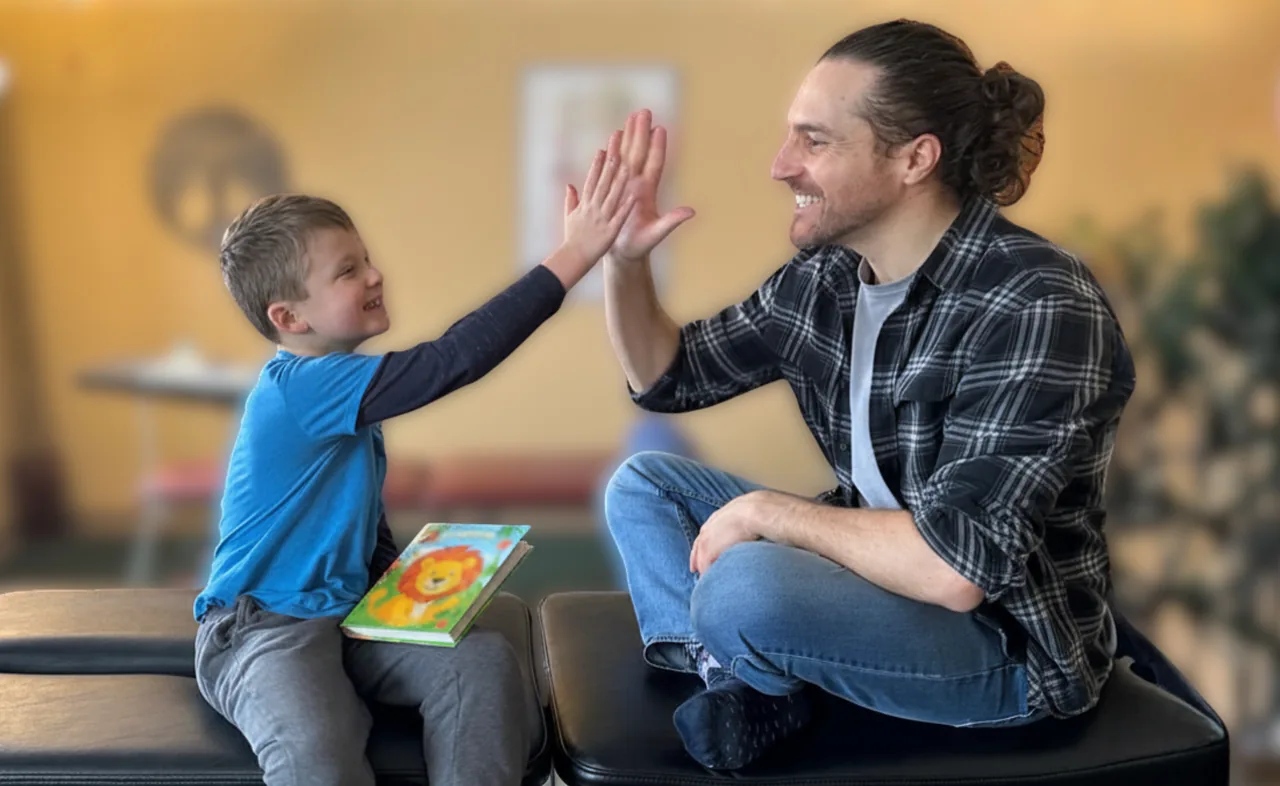 Dr. Zach Conner high-fiving a smiling young boy holding a lion picture book on the adjusting table at Van Every Family Chiropractic Center
